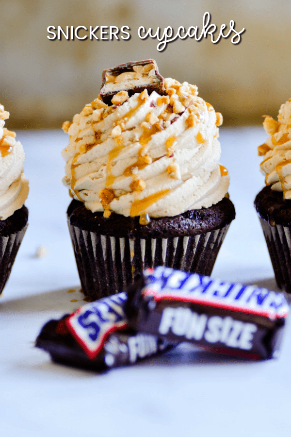 Fudgy chocolate Snickers Cupcakes piled high with peanut butter frosting and topped with caramel sauce, chopped nuts, and a candy bar.